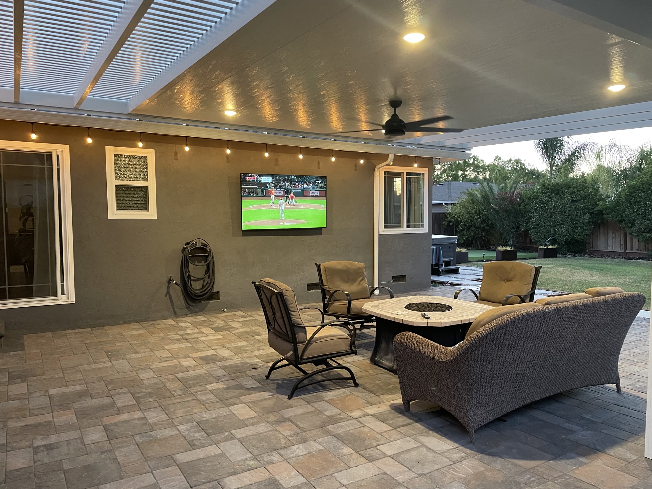 Finished covered patio with outdoor seating, ceiling fan, recessed lights, and wall-mounted TV by FG Pavers and Turf.
